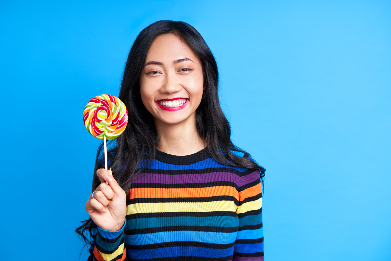 Woman wearing a colorful striped sweater smiling in front of a blue background holding a large rainbow lollipop and smiling.